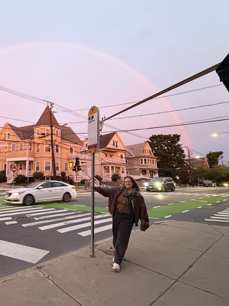 Miriam posing by a bus stop under a rainbow at sunset
